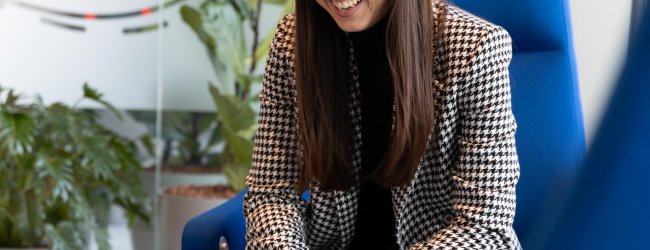 woman with brown hair sitting in a blue chair with laptop laughing